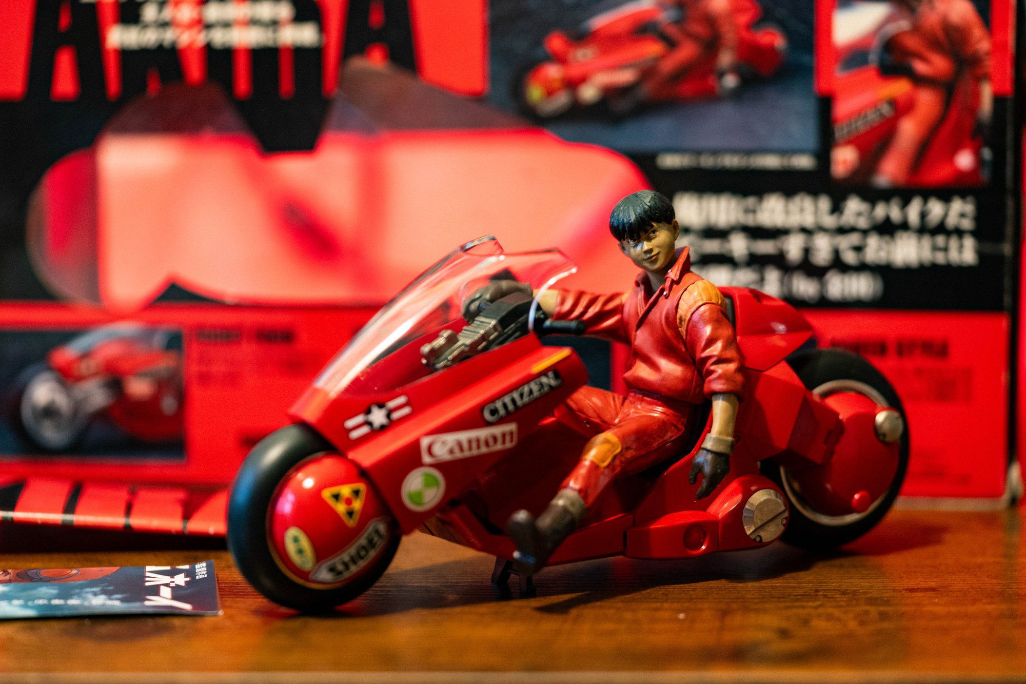 Bandai Soul of Popynica Kaneda figure seated on his red motorcycle, with Canon and Citizen markings and its box behind.